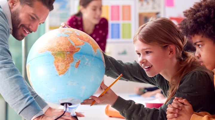 Teacher smiling as two students look at globe and one of them is pointing towards it