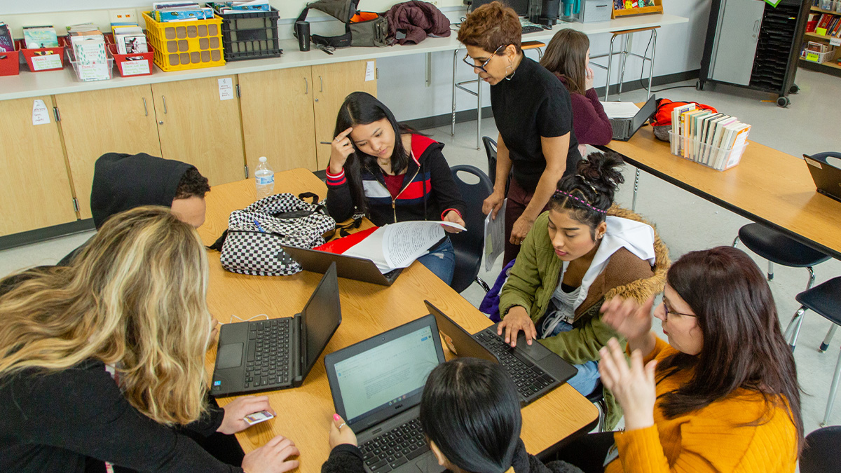 High school students sitting at a table with their laptops and teacher.