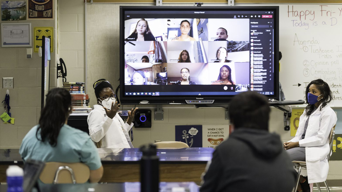 Teacher standing and two doctors sitting in front of a science classroom with students also participating remotely.