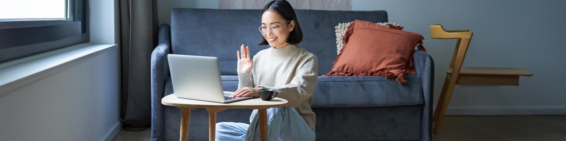 Student sitting in their living room smiling wand waiving at computer screen