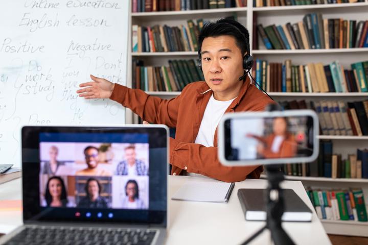 Teacher who is wearing headset and can be seen being filmed and on a Zoom meeting